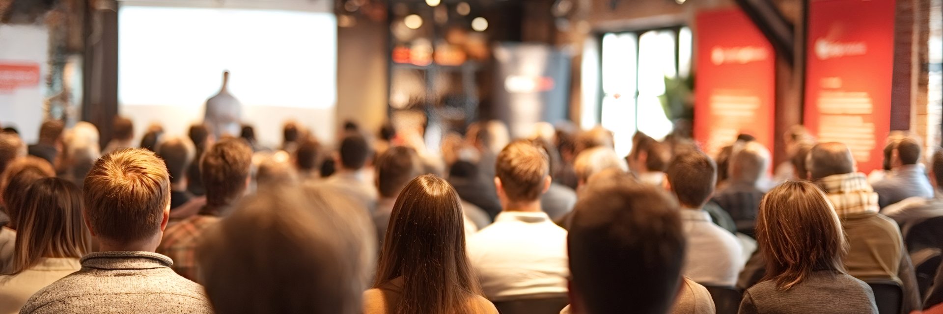 Une foule hétéroclite assiste à un séminaire éducatif ou à une présentation d'entreprise, les yeux rivés sur l'orateur sur scène, avec un écran flou en arrière-plan.