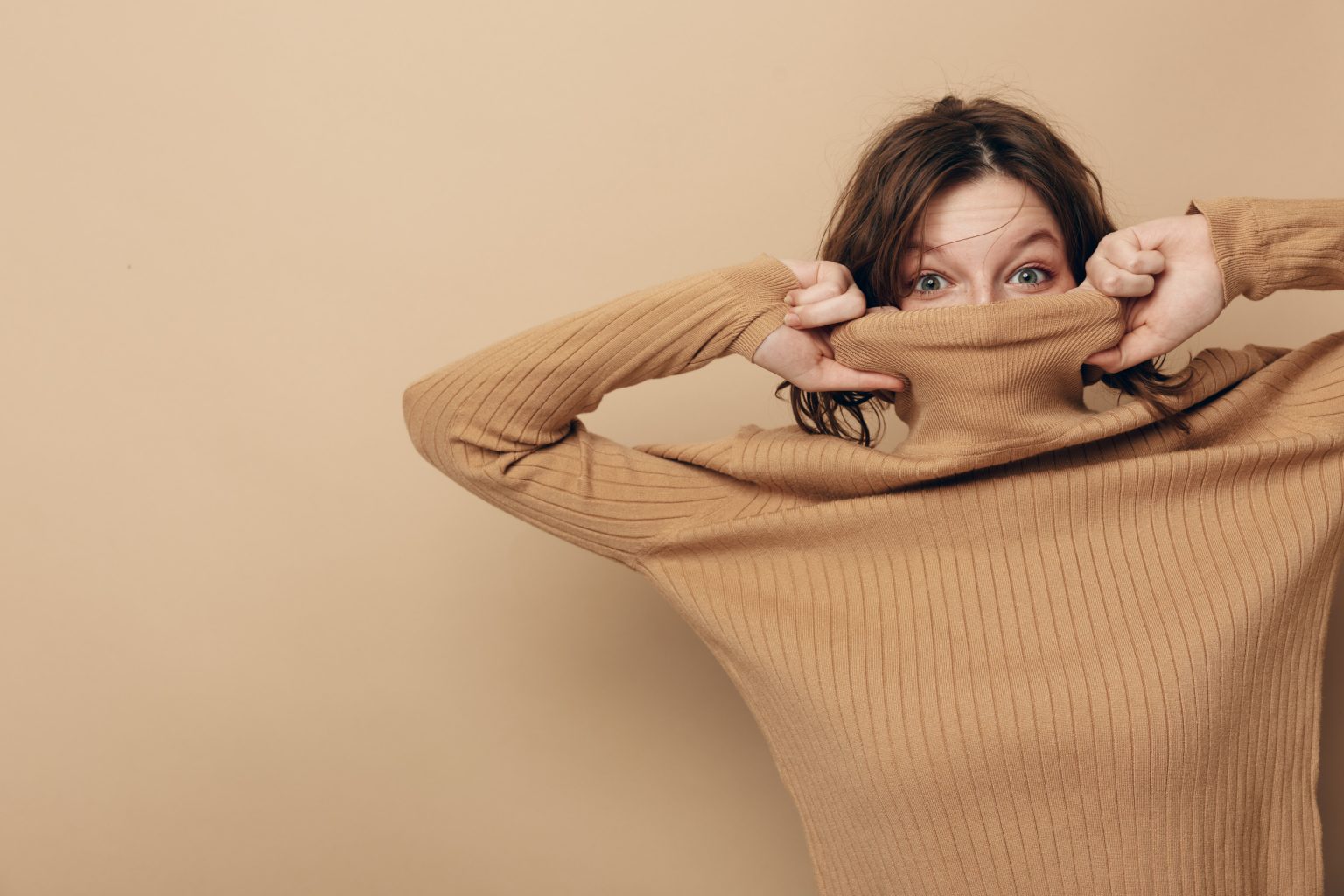 Jeune femme en pull marron tricoté, le col rabattu sur le visage, jette un regard en coin